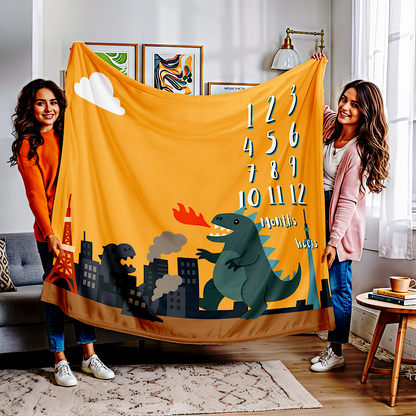 Two women holding a yellow kaiju-themed baby milestone blanket with numbers and a cityscape design in a living room.